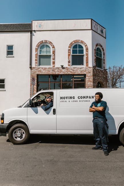 Two movers are engaged in a home relocation process beside a white van with its rear doors open, revealing stacked cardboard boxes inside. One mover, wearing a black puffer vest and a baseball cap, is
